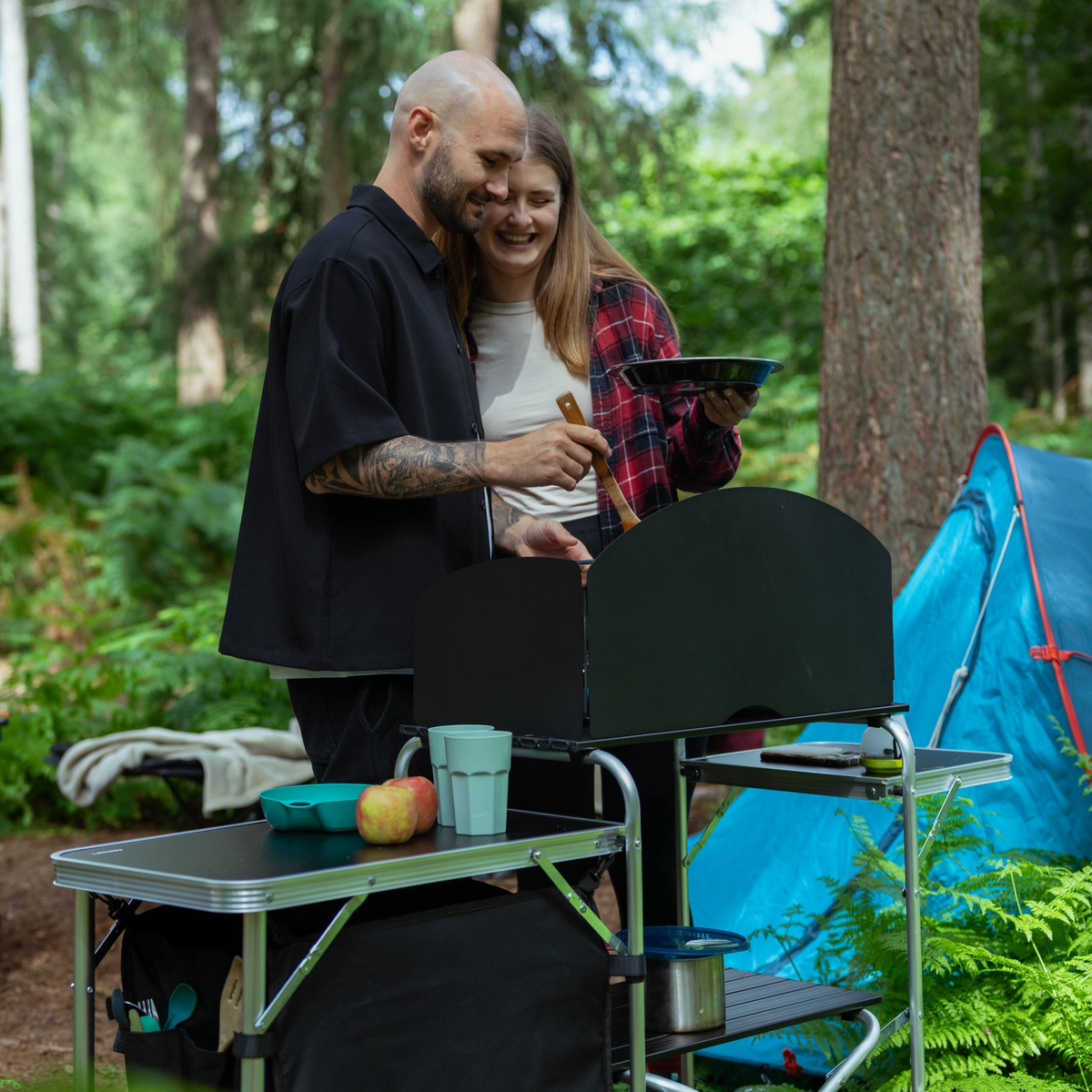 A couple cooking on a portable camping stove next to a blue tent in a forest.